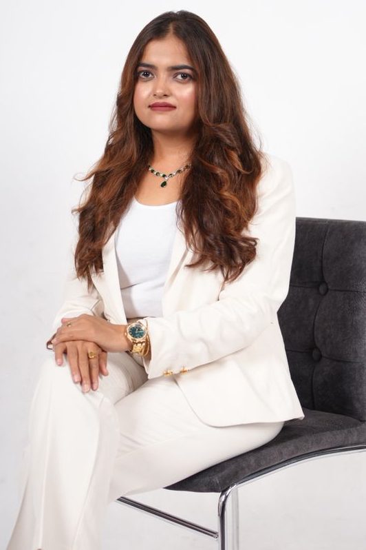 A woman in a formal white suit sits confidently on a chair with her arms crossed, featuring long hair and a stylish watch.